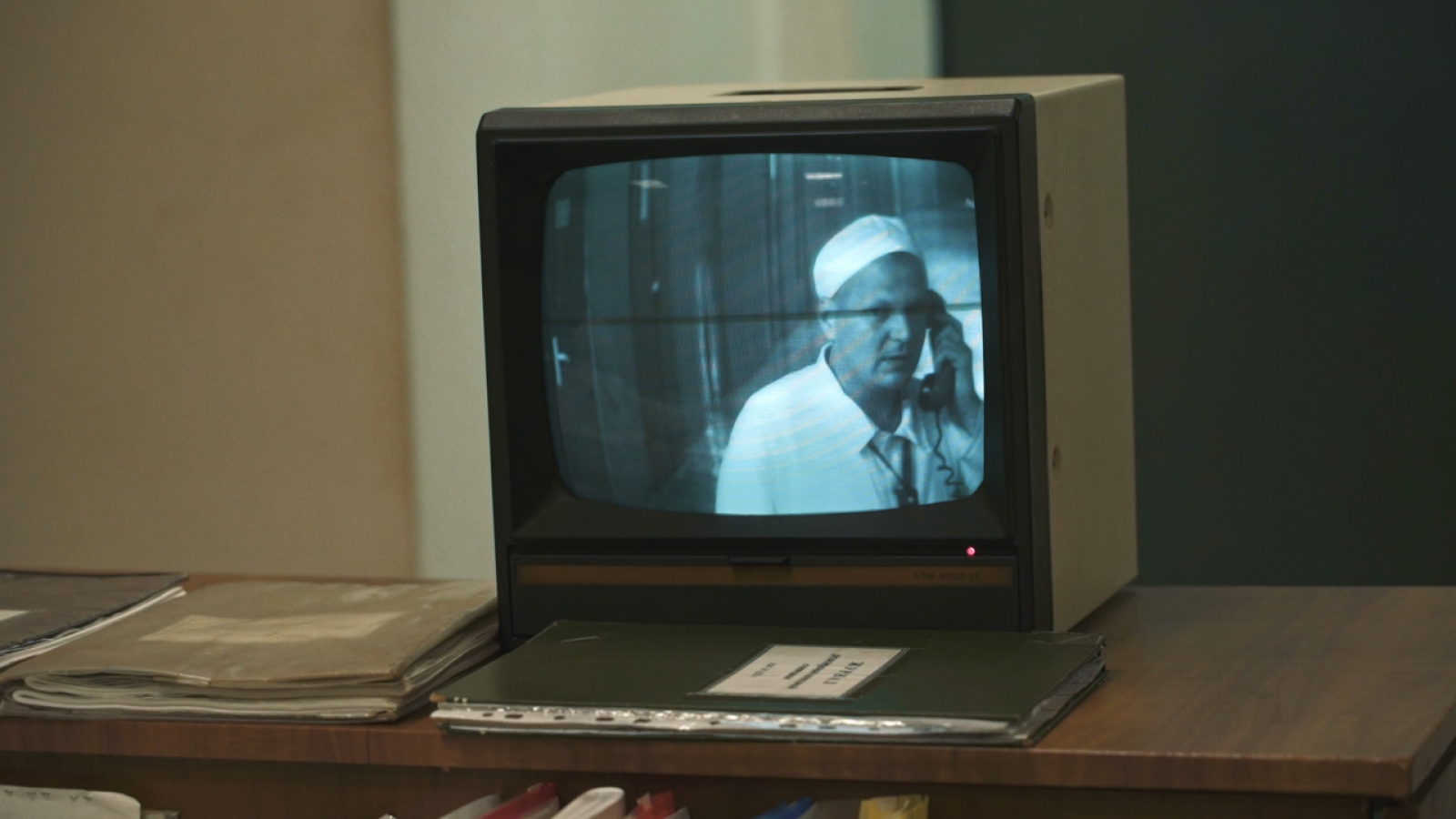 rus: На письмовому столі два зошити і телевізор. У телевізорі зображено, як працівник станції говорить по телефону.
