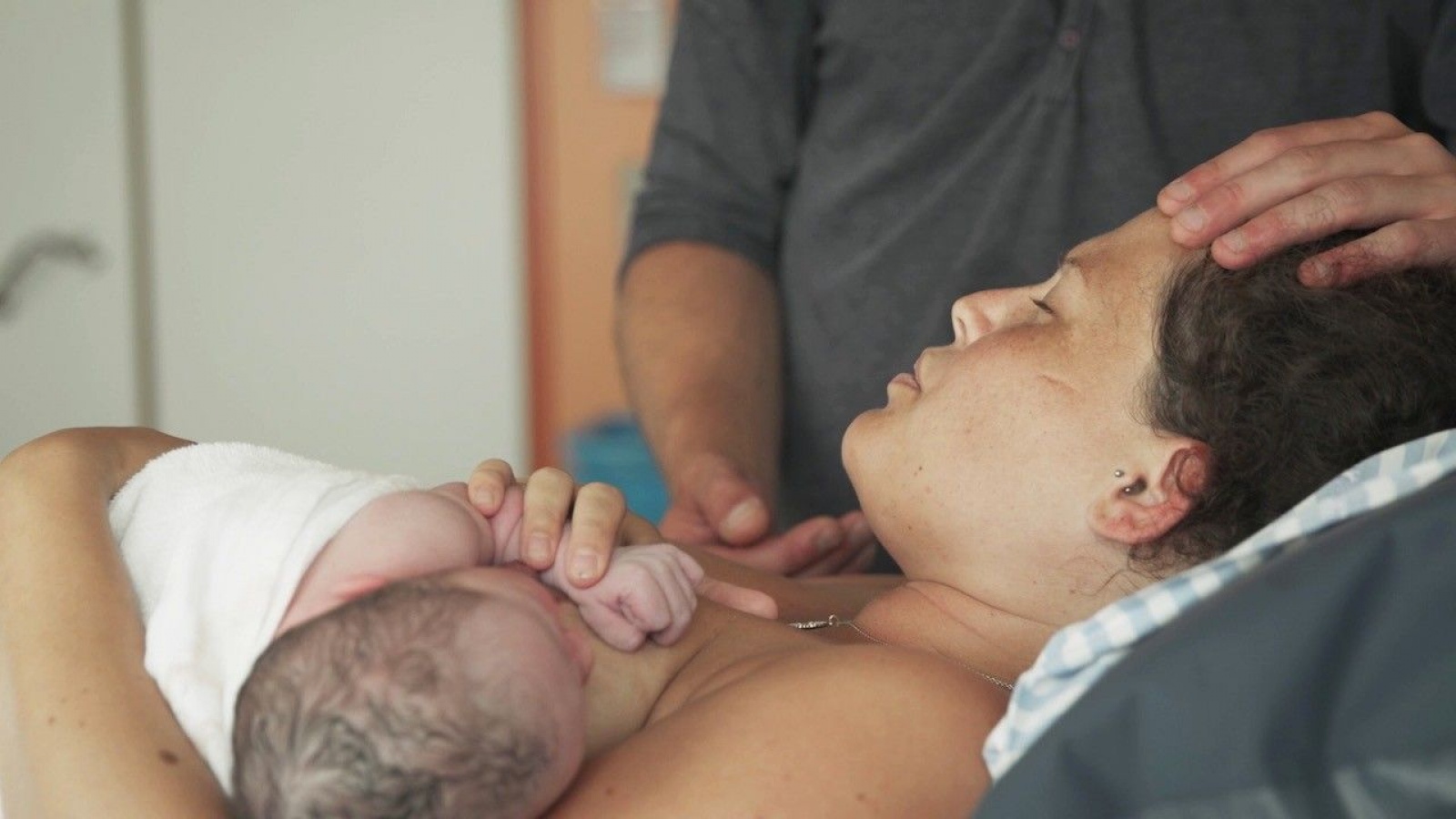 eng: Woman lying with a newborn on her chest. A man is standing nearby, putting his hand on her head.