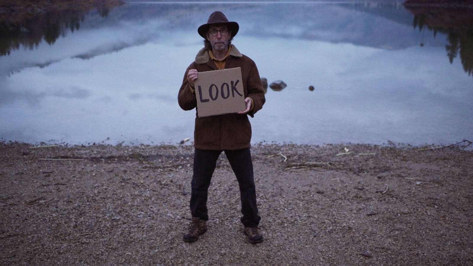 rus: Чоловік у капелюсі стоїть на березі озера з табличкою "Подивіться".