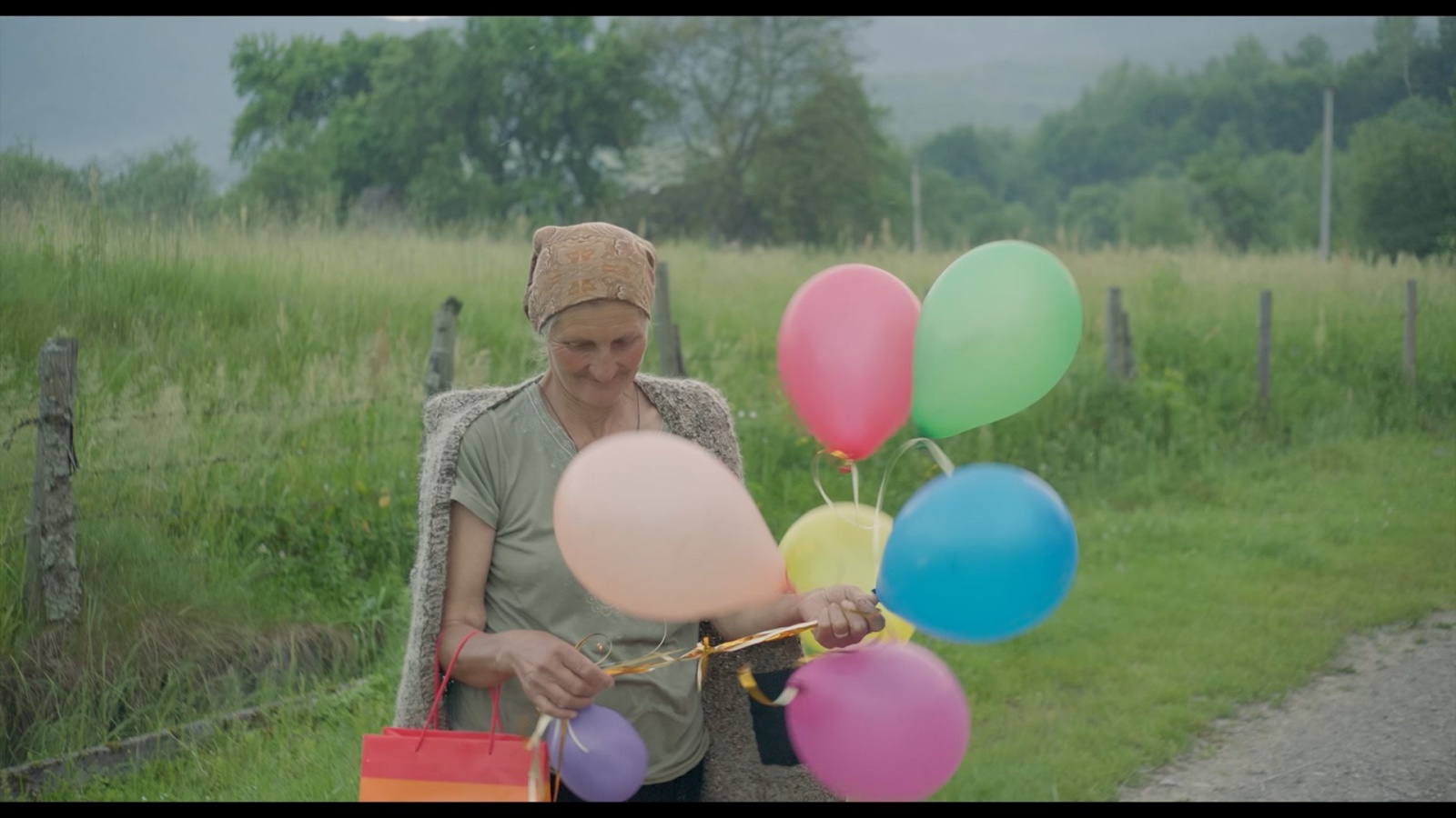 rus: Бабуся у селі тримає в руках різнокольорові кульки.