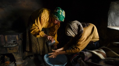 Дві літні жінки сидять у напівтемряві, вбрані у жовтий одяг. Жінка у зеленій хустці миє руки жінці у коричневій хустці.
