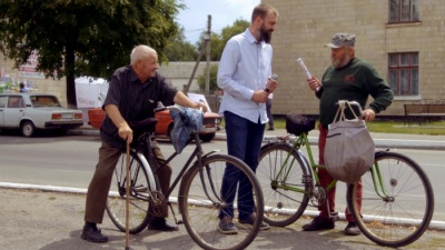 three men, the man in the center in a shirt, two men with bicycles at the edge