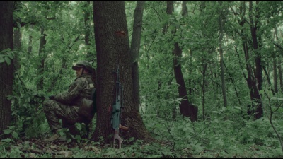Військовий у лісі навприсядки спирається на дерево. Поряд із деревом автомат військового.