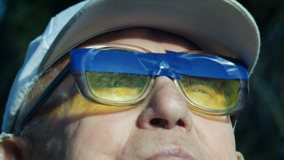 Old man looks through yellow glasses covered with a blue patch.